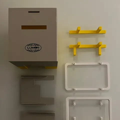 Flat lay of 3D-printed prototype parts: a grey Lumon-branded printer enclosure, yellow printer cradles, white display frames with mounting brackets, grey side panels and small brackets